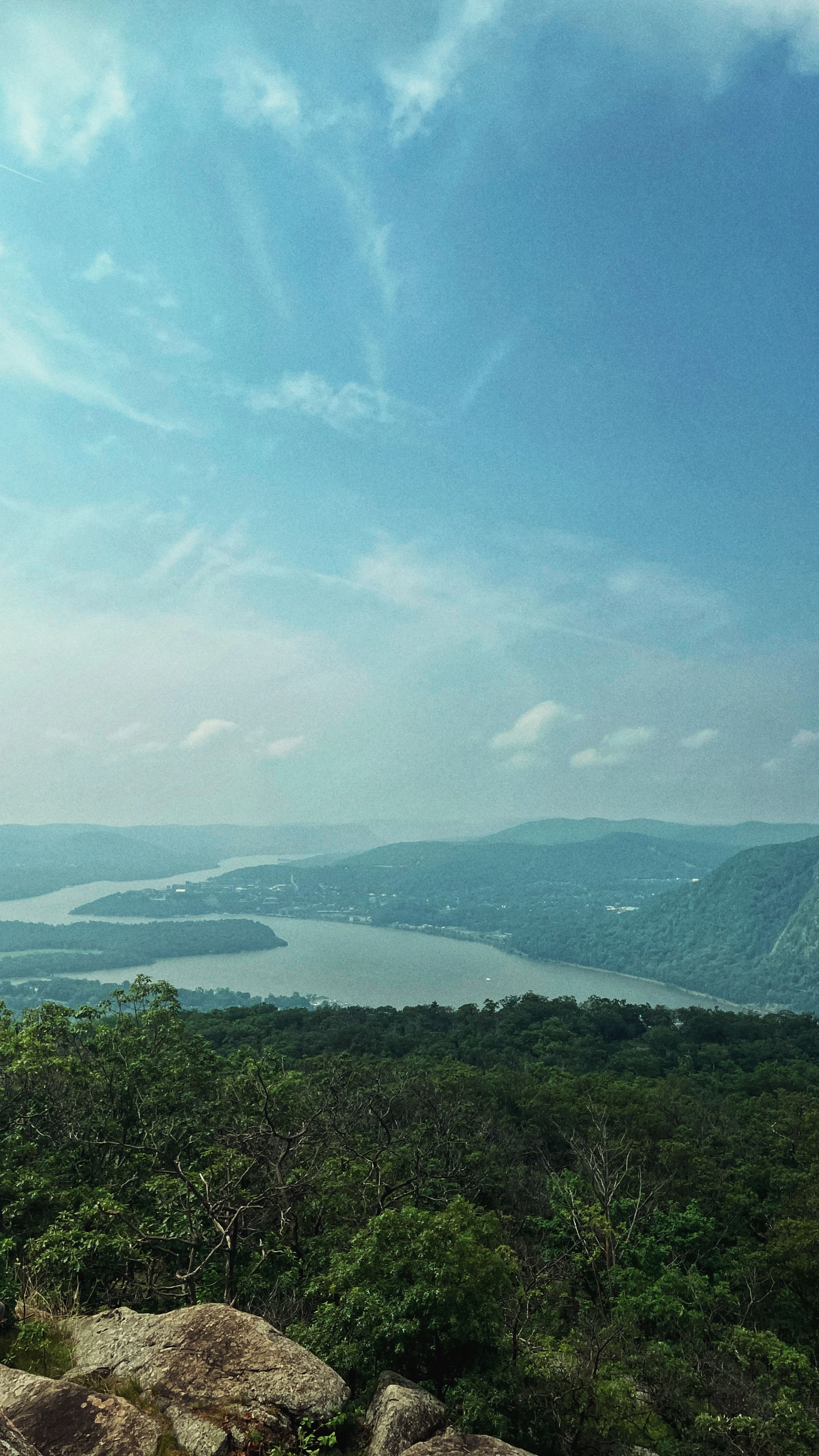 Sweeping view over a forested river valley from a rocky overlook, a wide bend of water framed by hazy ridgelines under a pale blue sky