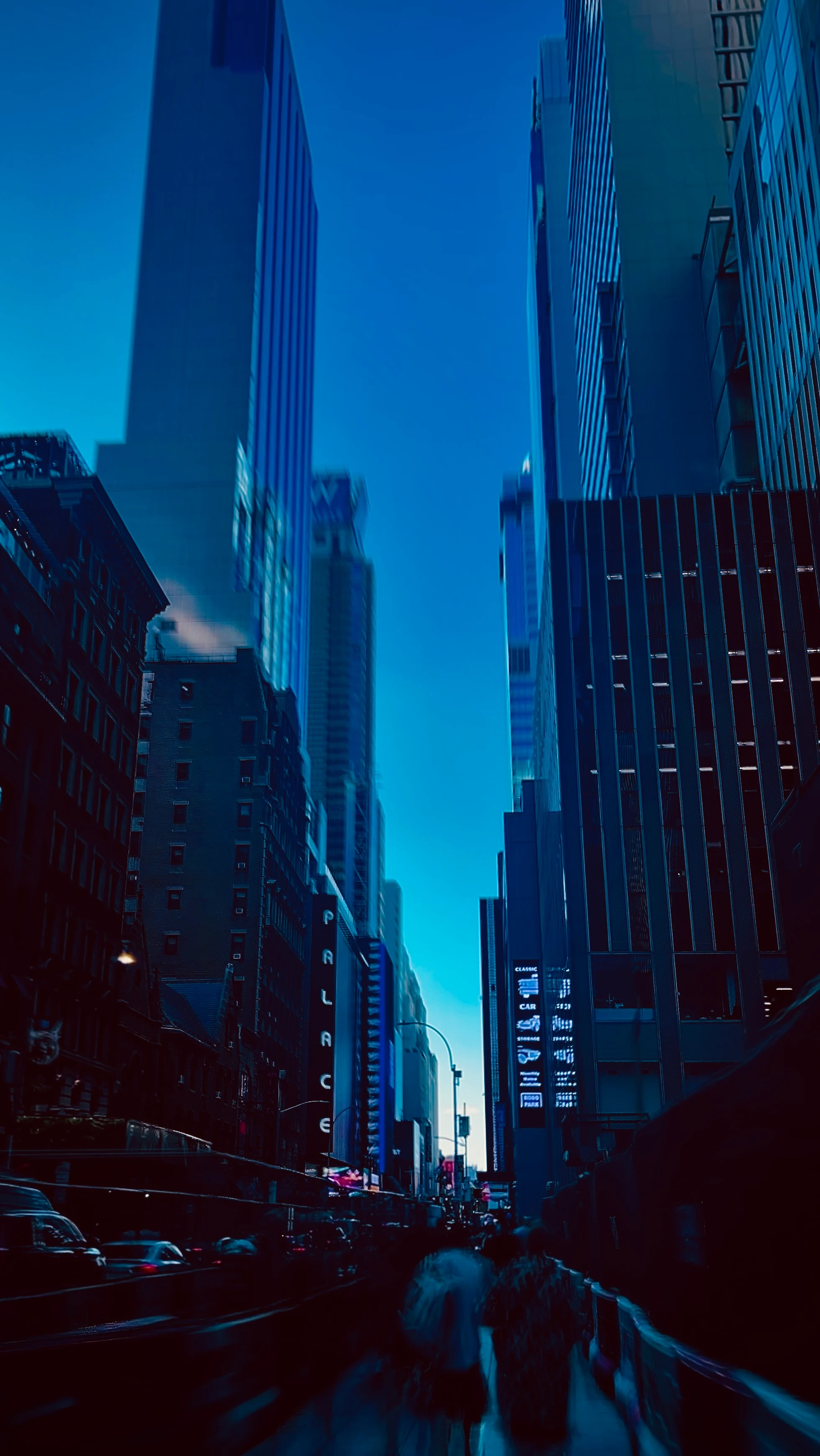 Twilight view up a dense city street between illuminated skyscrapers, pedestrians and taxis blurred in motion at street level