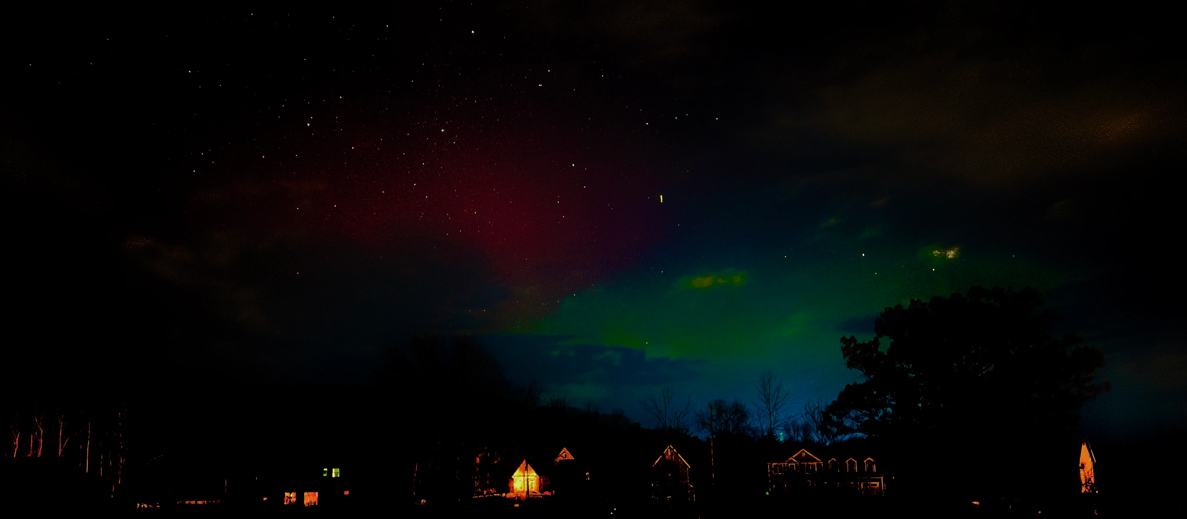 Wide night sky panorama of red and green aurora borealis glowing above a starlit horizon of illuminated houses