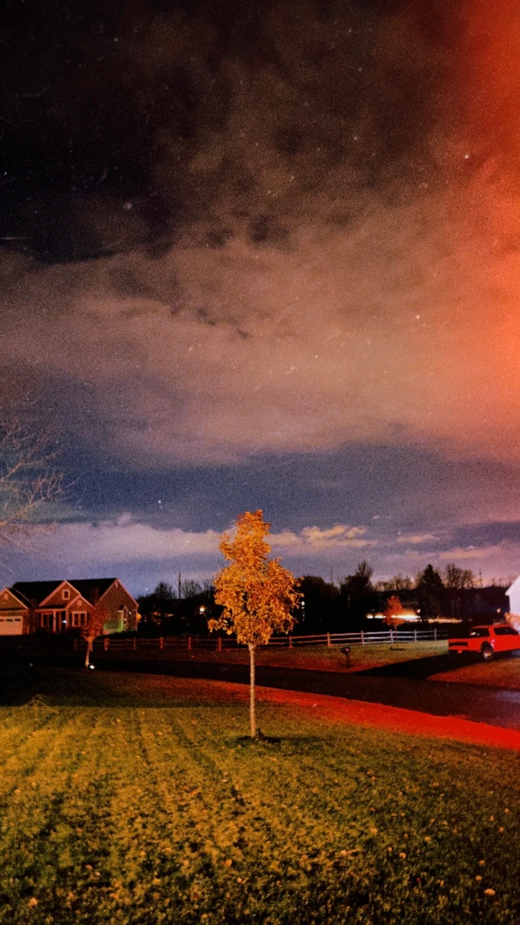 A lone illuminated tree on a suburban lawn at night beneath a dramatic sky glowing orange and blue from storm light