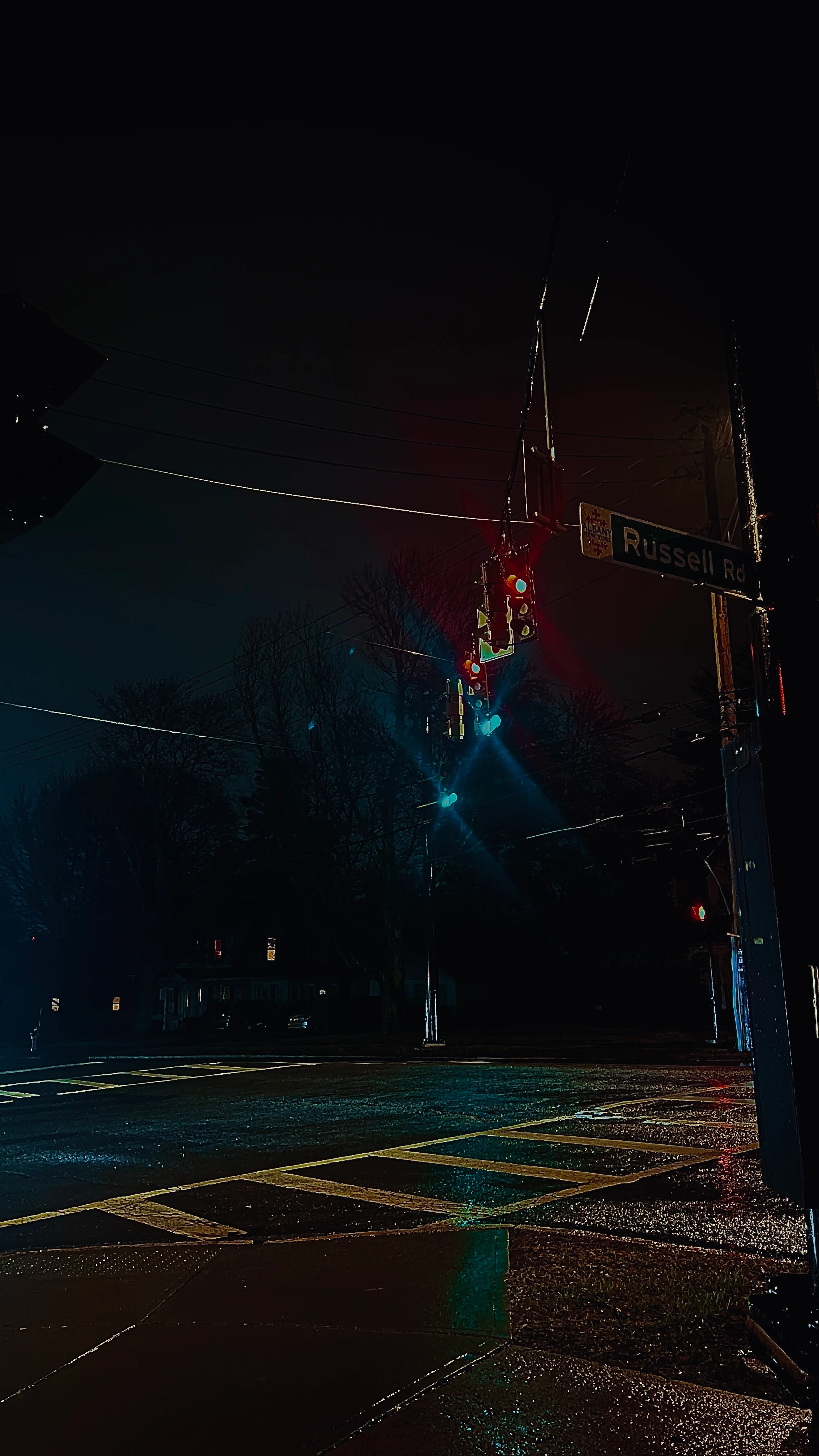 Empty night street intersection with a glowing traffic signal and 'Russell Rd' street sign, rain-slicked crosswalk reflecting light