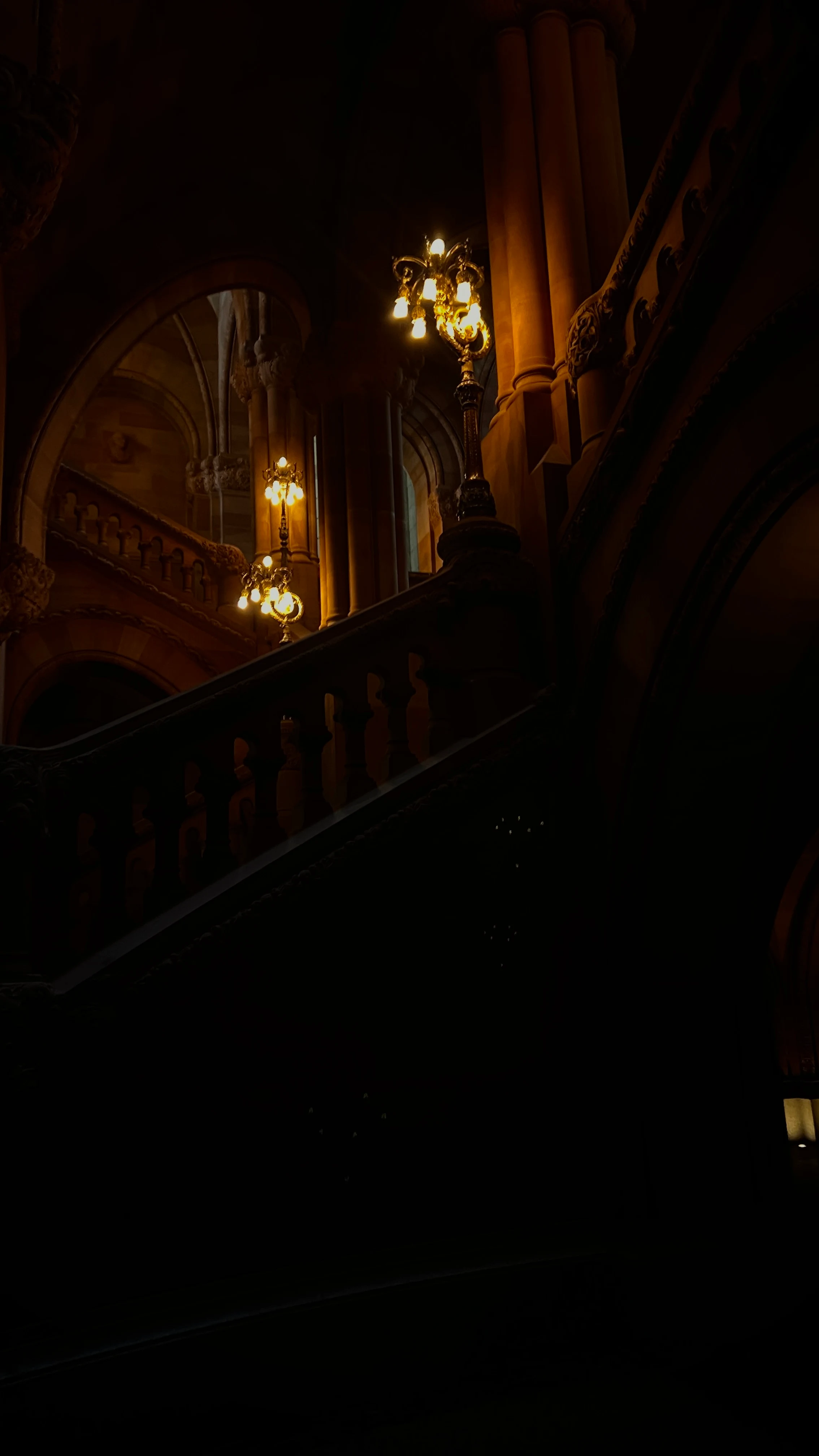 Dark gothic stone stairwell lit by two ornate multi-bulb lamps, arched stonework and columns rising into shadow behind the banister