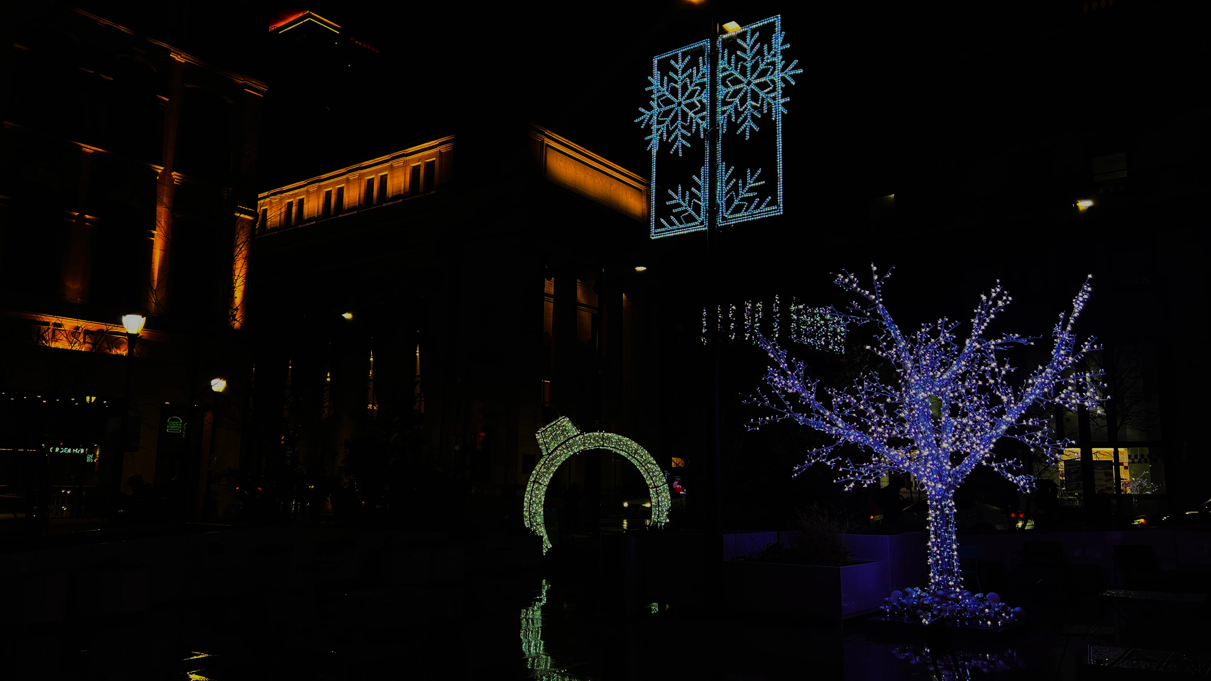 City plaza at night decorated with illuminated holiday lights, a glowing tree, snowflake light sculpture, and an arch beside historic buildings