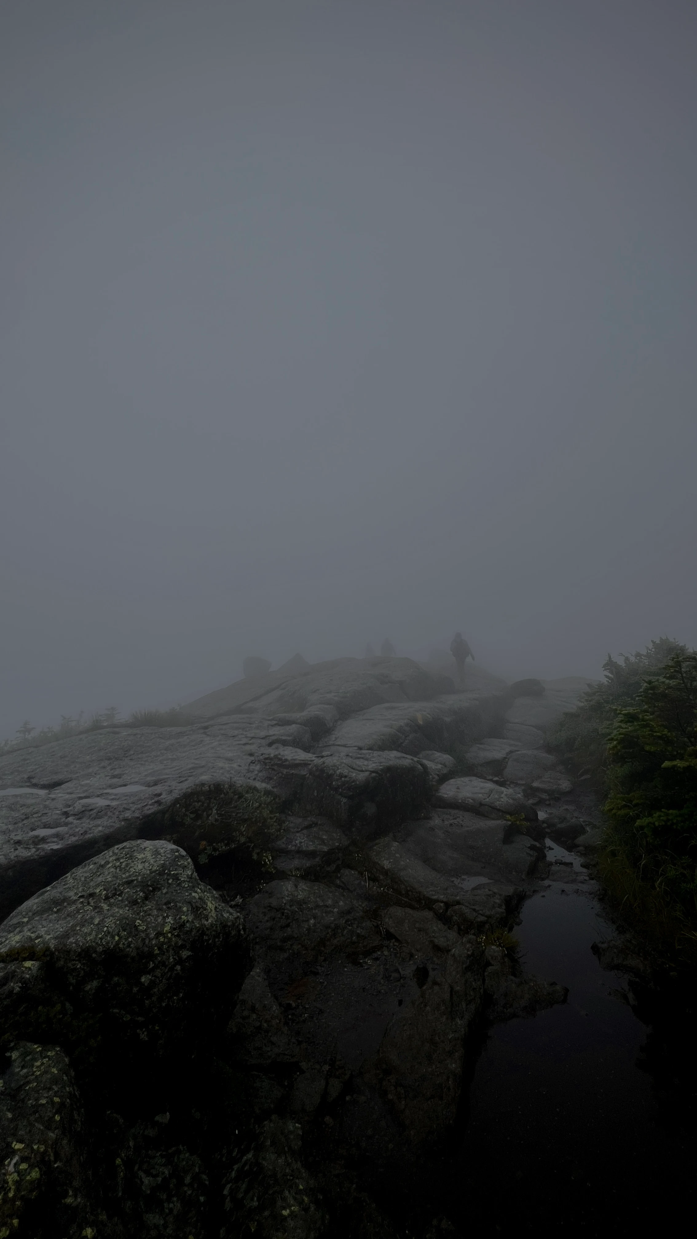 Two hikers walking along a rocky ridge trail engulfed in thick fog, visibility reduced to silhouettes