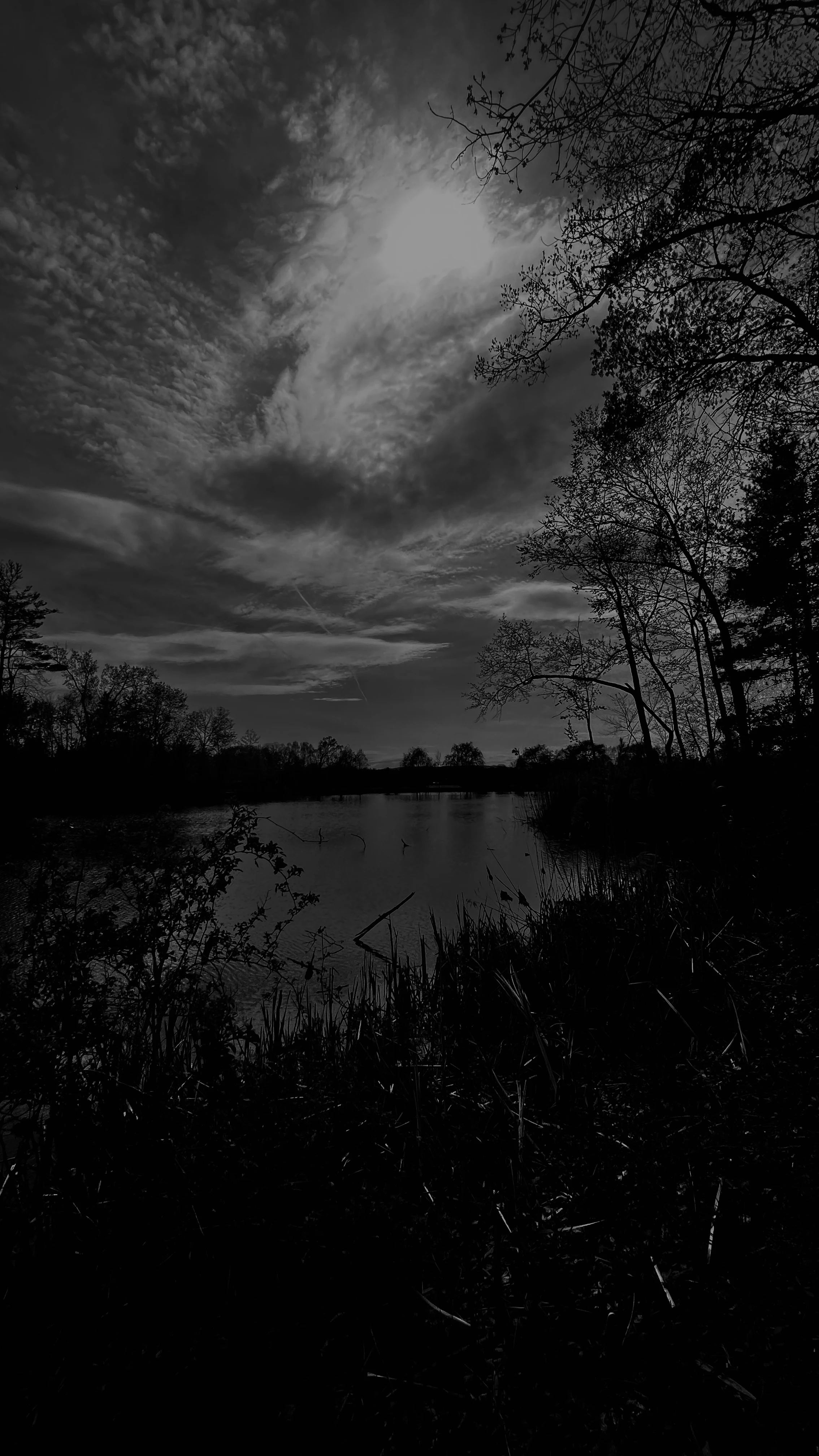 Black-and-white view of a calm lake beneath a dramatic sky, sunlight breaking through streaked clouds above a silhouetted shoreline