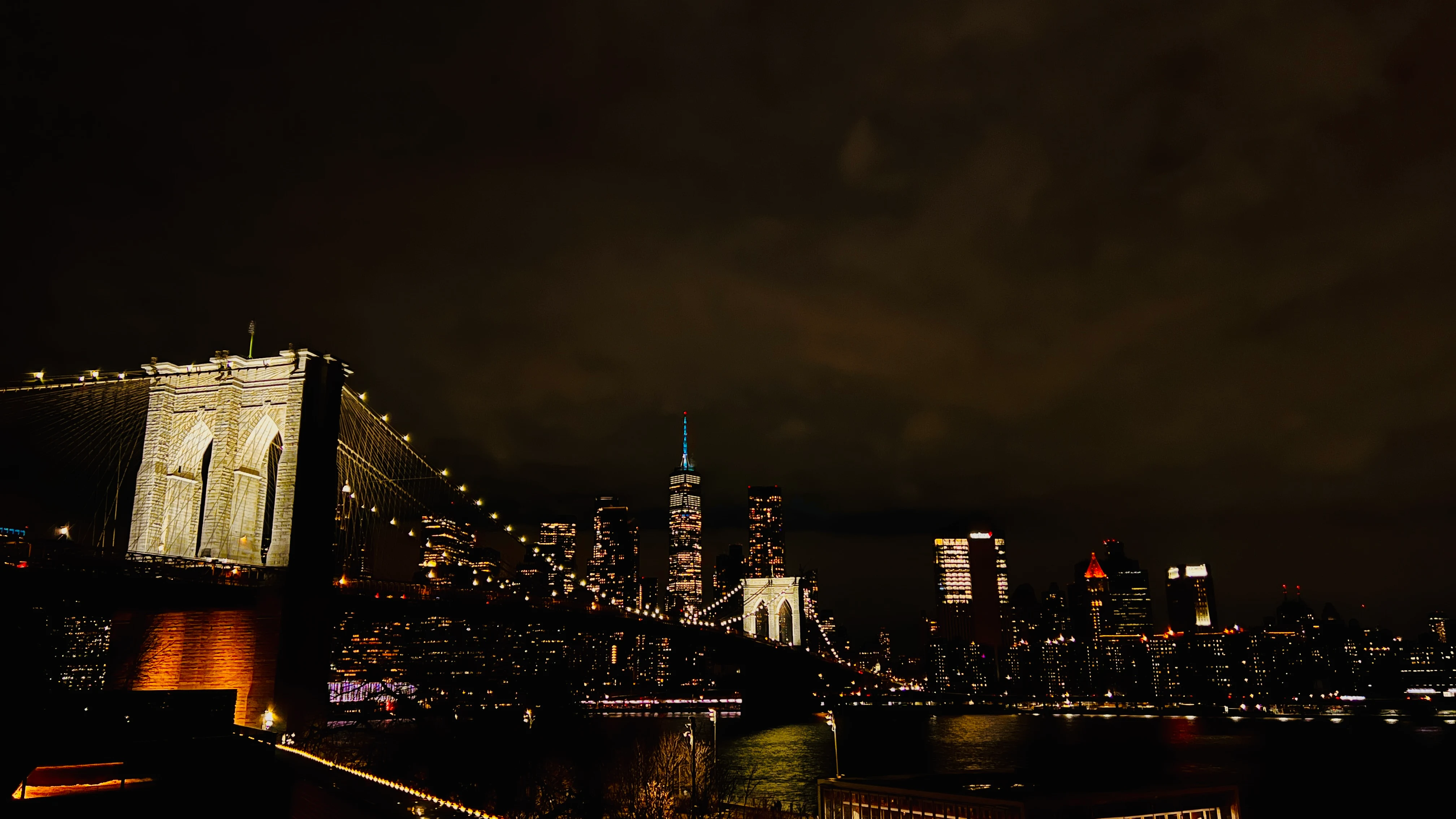 Brooklyn Bridge and the illuminated Manhattan skyline at night, reflections shimmering on the water below