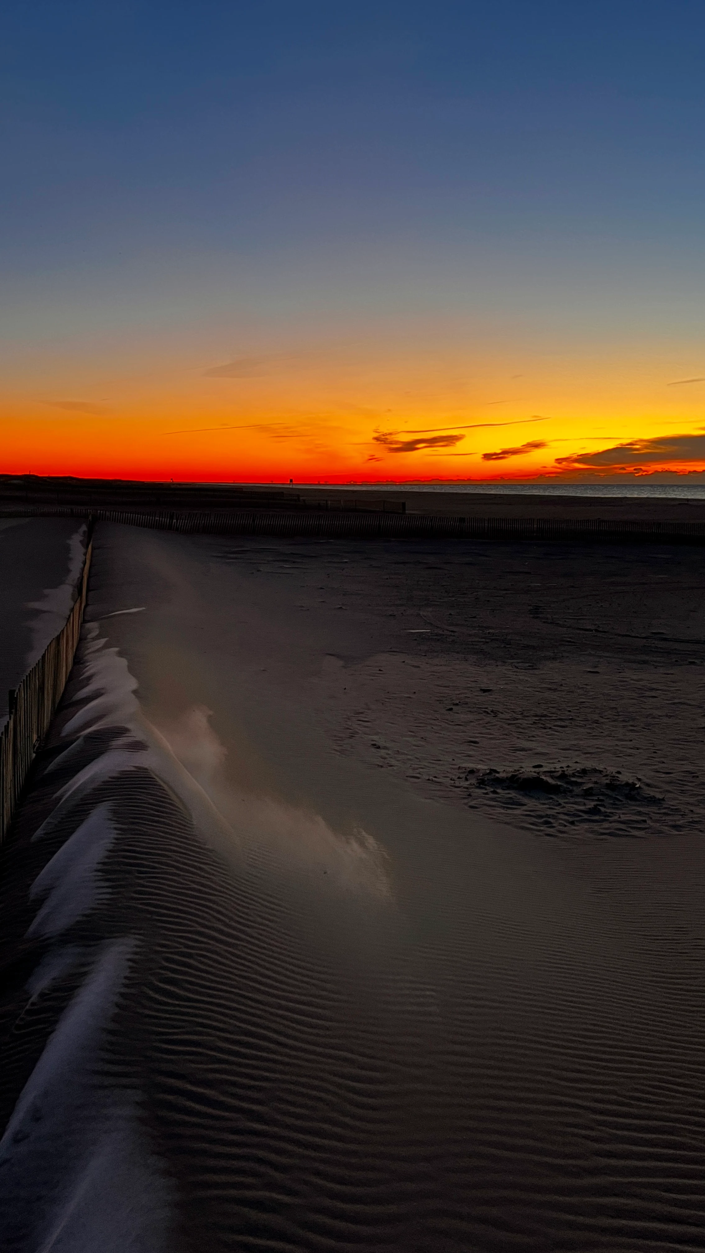 Wind-rippled sand dunes along a beach at dusk, a wooden fence line leading toward an orange horizon over the ocean