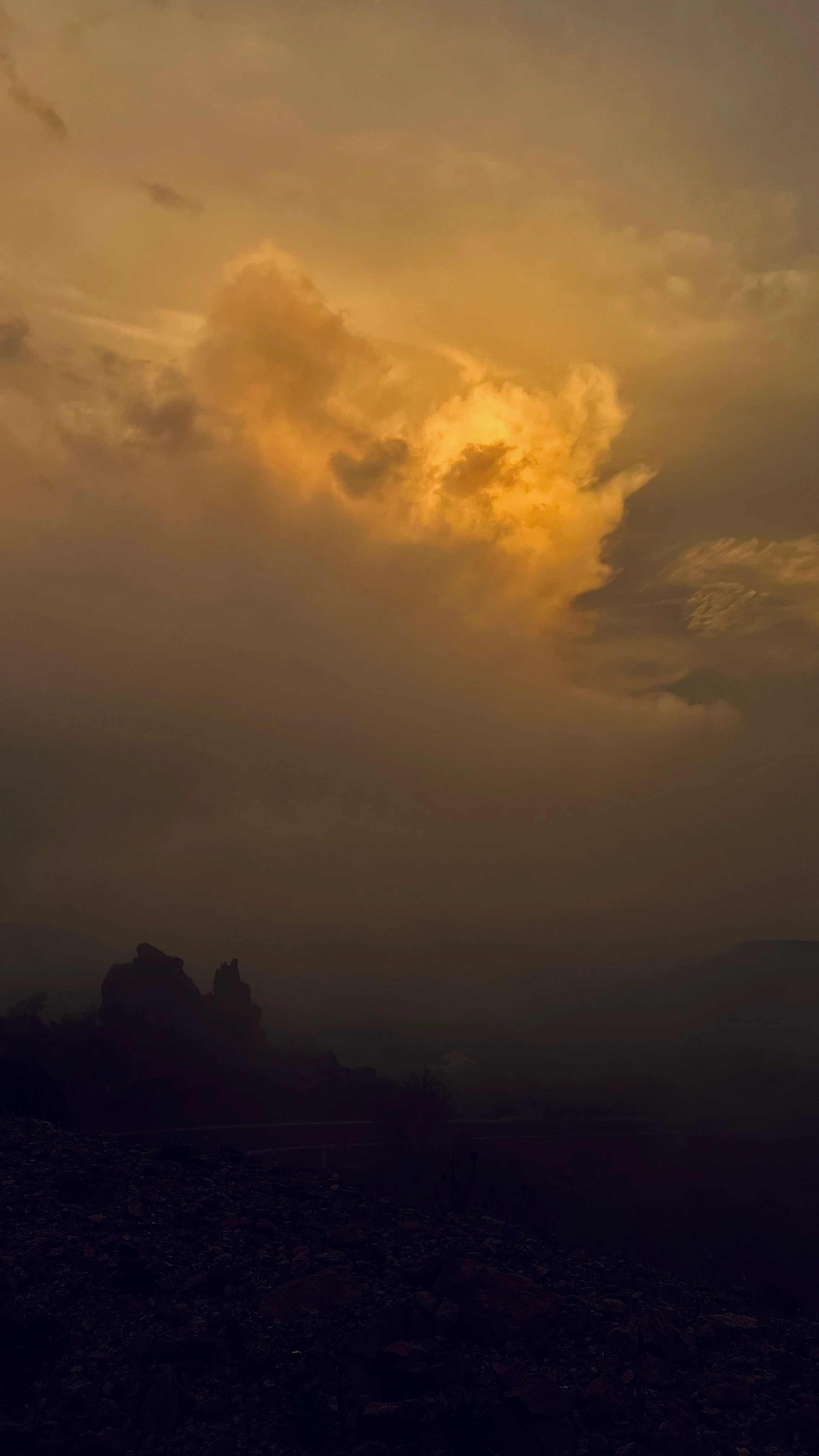 Golden storm clouds glowing at sunset over silhouetted desert mountains and rocky foreground terrain