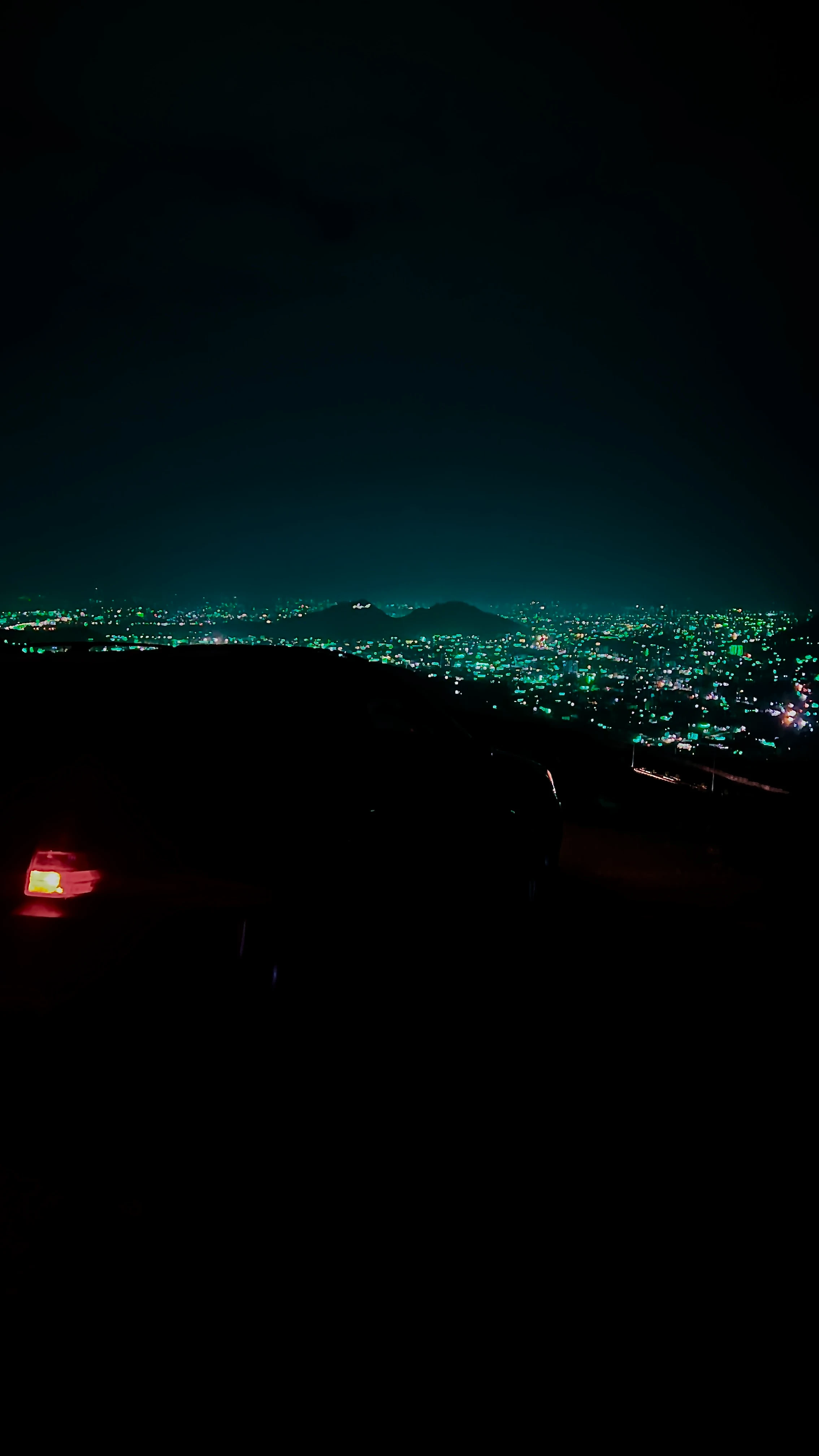 Aerial night view of a city's teal-lit grid from a dark hillside, a car's red tail light glowing in the foreground