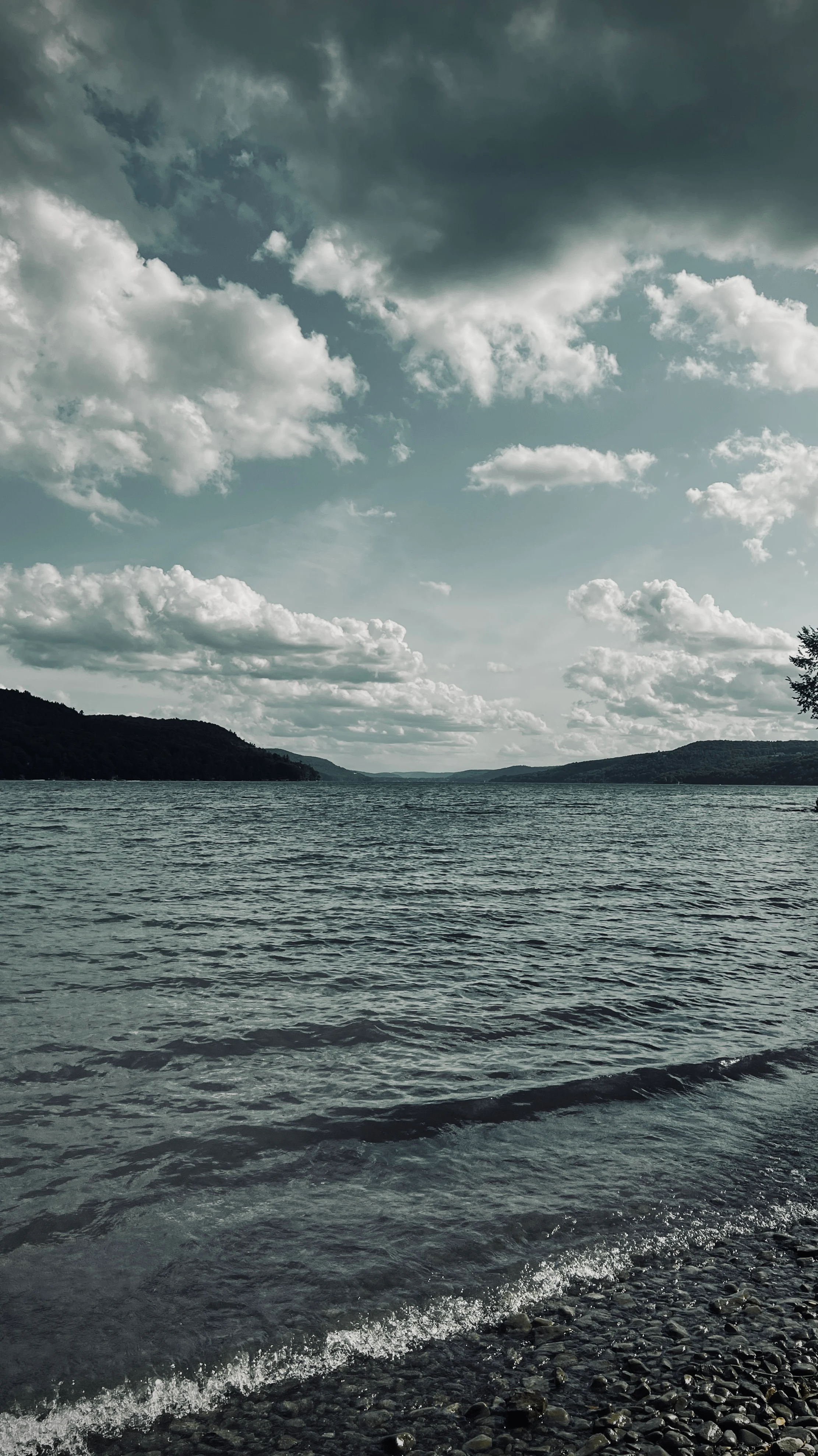 Calm lake shoreline with a pebble beach in the foreground, rippling water stretching toward tree-lined hills under a cloudy sky