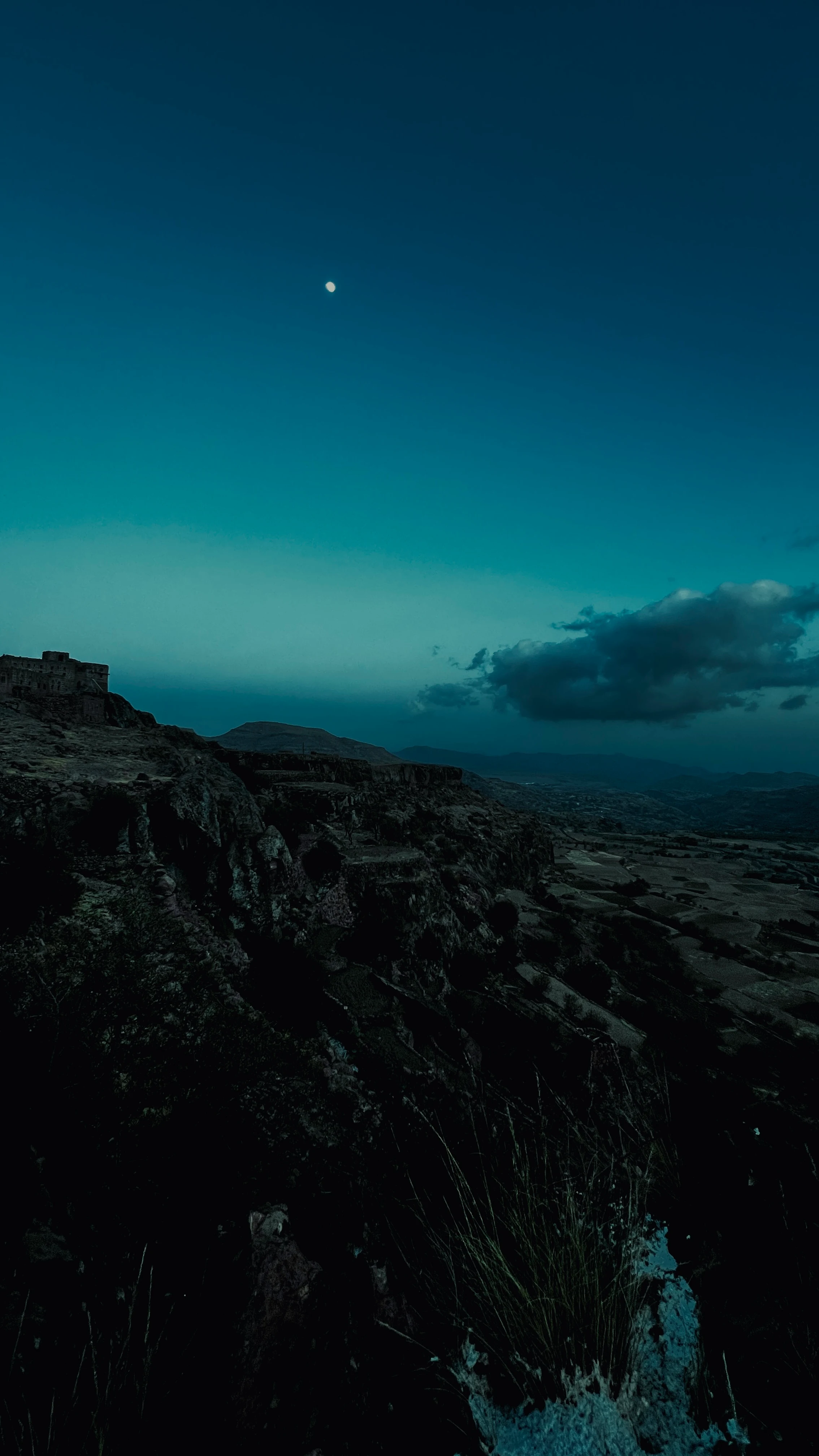 Dusk view from a cliff edge over a silhouetted fortress ruin and rolling valley, a pale moon rising in the blue sky
