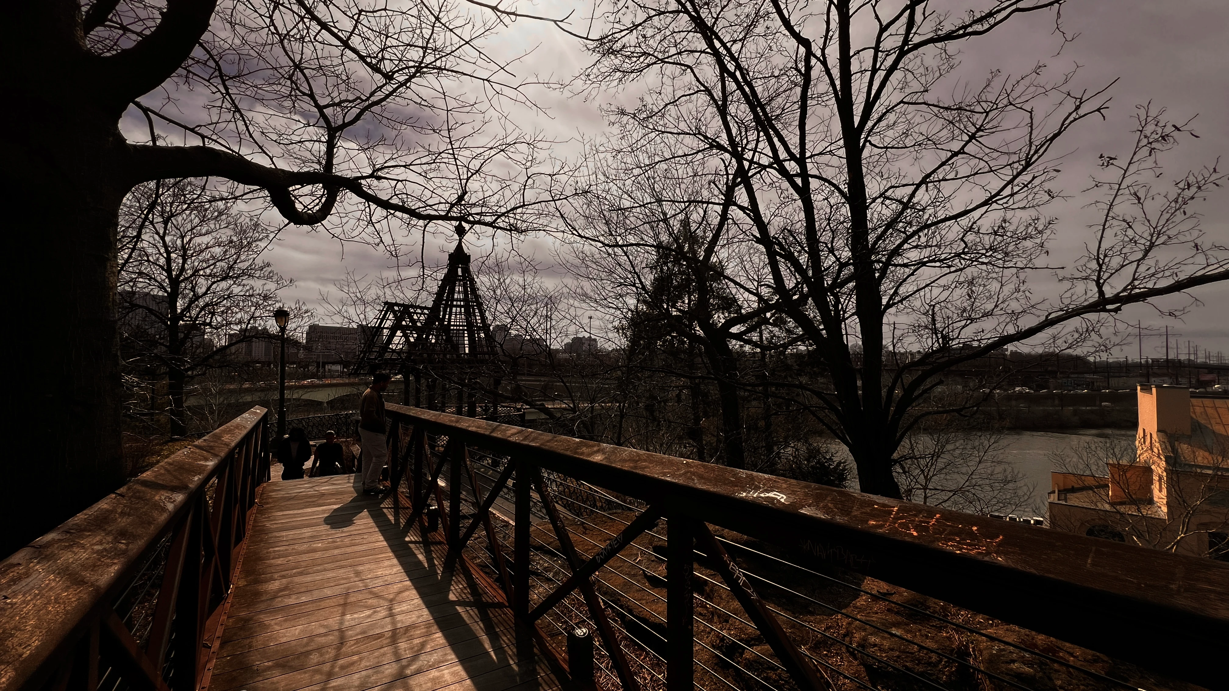 Wooden boardwalk winding through bare winter trees above a river, a city skyline visible in the hazy distance