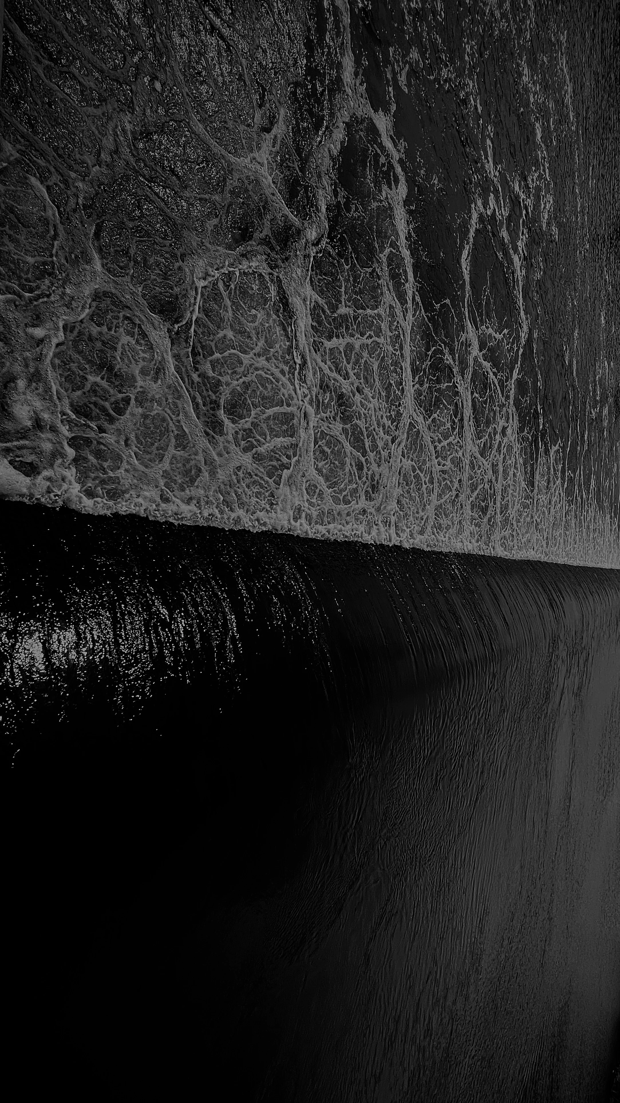 Close-up black-and-white abstract of a breaking ocean wave, lace-like foam patterns against dark water